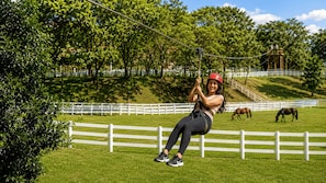 A person zip-lining over a grassy area with white fences and trees in the background. - Hotel Fazenda Águas de Lindóia (Águas de Lindóia)