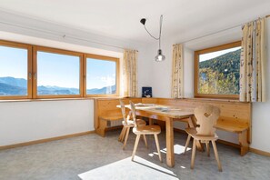 A room with a large wooden table, two chairs, a bench, and a view of mountains through large windows.