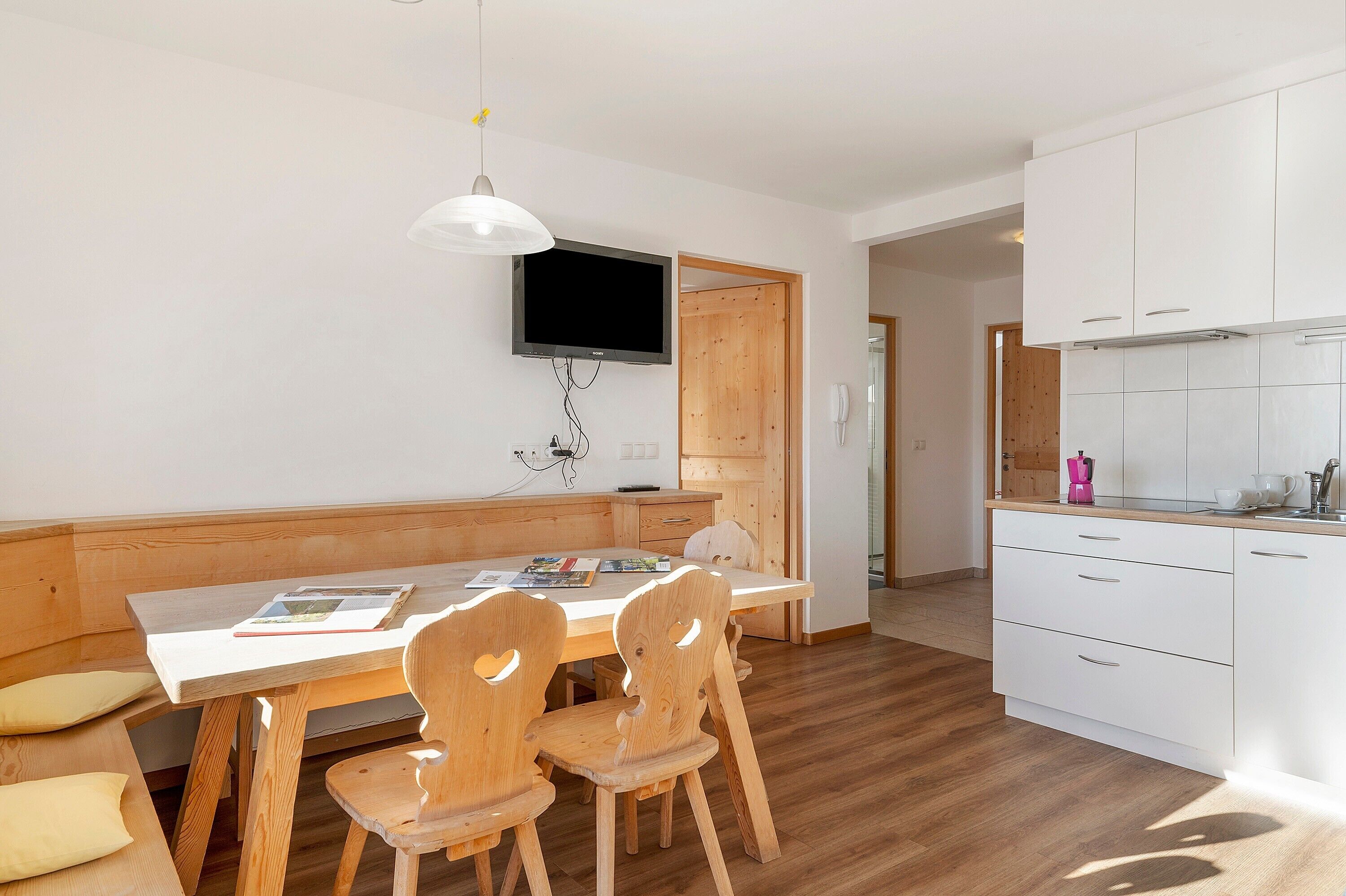 A modern kitchen with white cabinets, a wooden dining table with chairs, and a TV mounted on the wall.