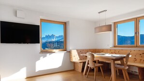 Une pièce avec un téléviseur fixé au mur, une table à manger avec des chaises, un banc et de grandes fenêtres donnant sur la montagne.