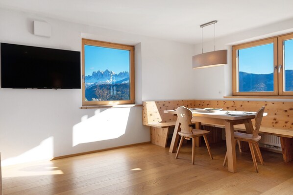 Une pièce avec un téléviseur fixé au mur, une table à manger avec des chaises, un banc et de grandes fenêtres donnant sur la montagne.