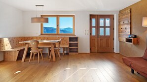 Une pièce avec un bureau en bois, des chaises, un luminaire suspendu, une porte en bois et une vue sur les montagnes par une grande fenêtre.