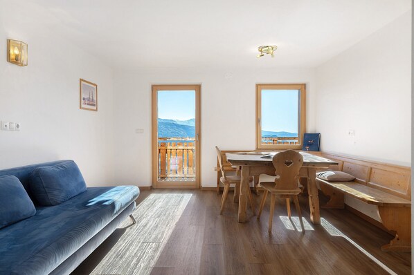 A room with a sofa, wooden dining table, chairs, a bench, and a view through a door.