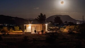 Una piccola costruzione illuminata con una porta e finestre, circondata da vegetazione rada e colline sullo sfondo.