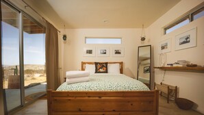 Una camera da letto con un letto grande, una porta scorrevole in vetro che dà su una vista del deserto, una piccola finestra a lucernario e un comodino con dei libri.