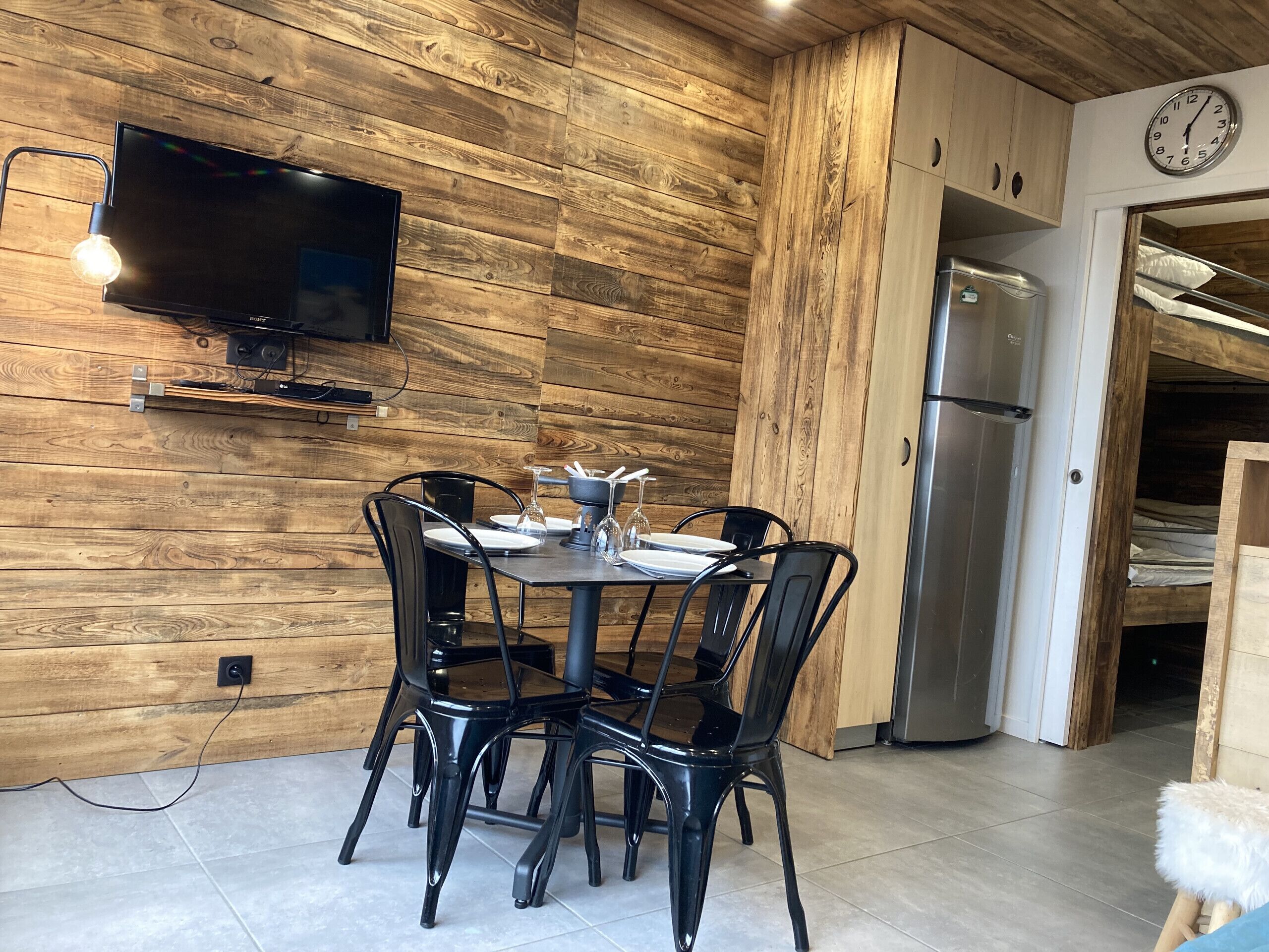 A modern kitchen with wooden walls, a dining table set for four, a built-in TV, and a refrigerator.