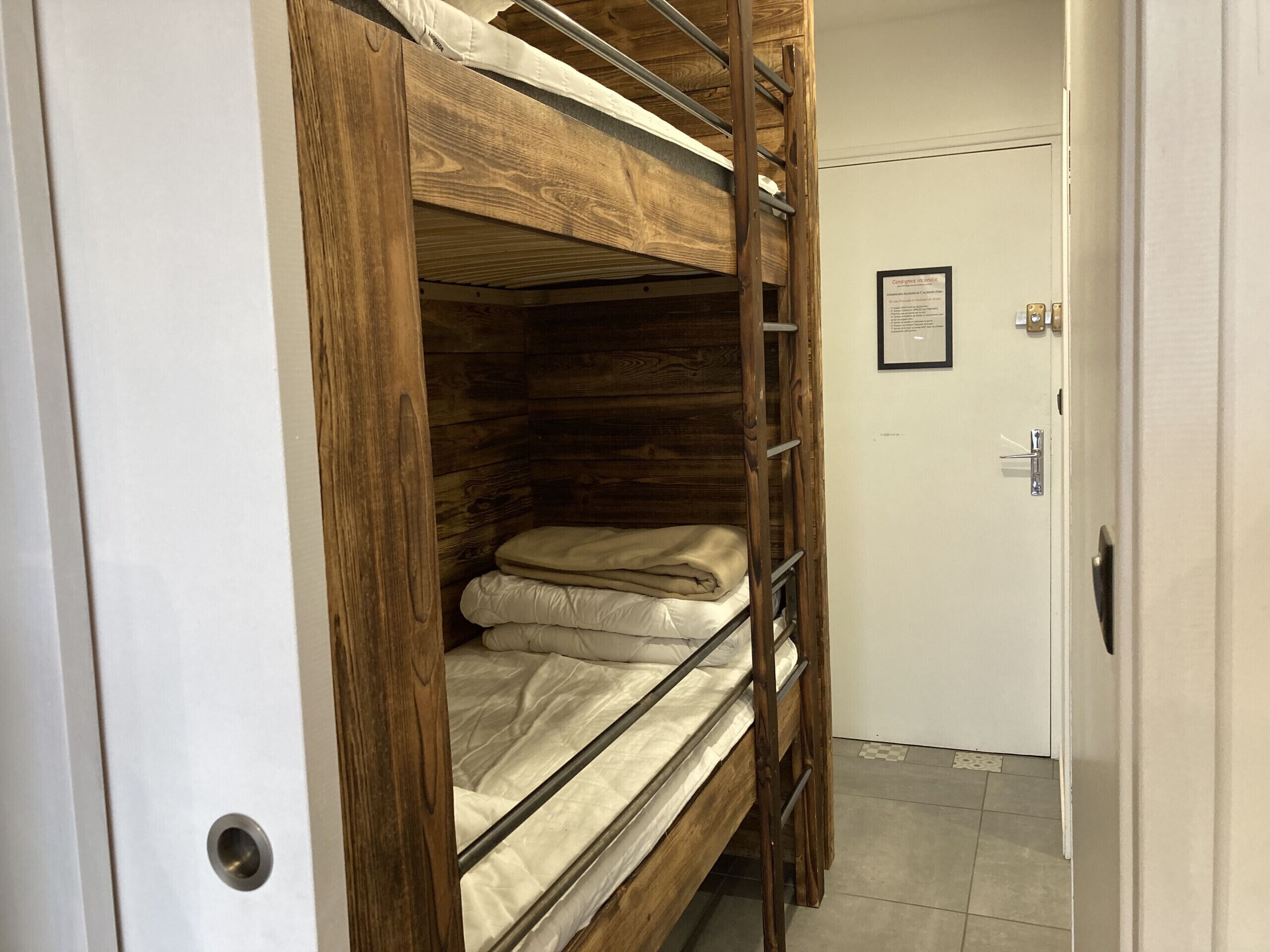 A bunk bed with a wooden frame and metal ladder, a white door, and a framed notice on the wall.