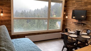A cozy room with a large window, a bed, a dining table with chairs, and a mounted TV.