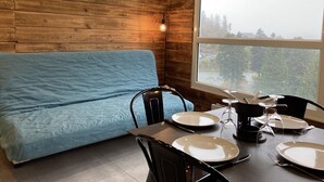A room with a wooden wall, a large window, a blue sofa, and a dining table set with plates and glasses.