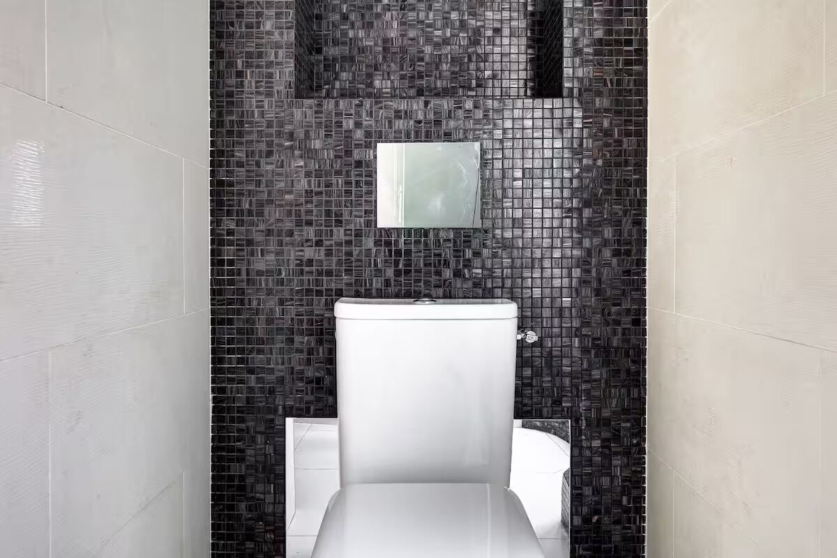 Ein modernes Badezimmer mit einer weißen Toilette, einer Wand mit Mosaikfliesen und einem kleinen Spiegel über der Toilette.