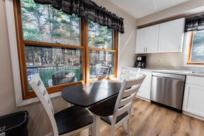A dining area with a table and chairs, a kitchen with white cabinets and stainless steel appliances, and a view of trees through large windows. - Kavanagh Bay Resort|Hot Tub&Sauna #1 (Hayward)