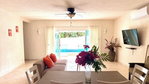 A dining area with a table, chairs, a sofa, a ceiling fan, a TV, and a view of an outdoor swimming pool. - Marvelous 3-bedroom Apartment in Villa, Beautiful Pool, Steps from Surf & Ocean (Atlantic Shores)