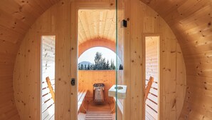 A wooden interior with a central staircase leading to a balcony, a window with a view of trees and mountains, and a small outdoor seating area.
