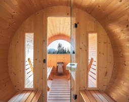 Un interior de madera con una escalera central que conduce a un balcón, una ventana con vistas a árboles y montañas, y un pequeño espacio exterior con asientos.