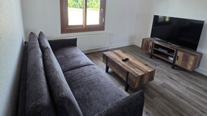 En moderne stue med en hjørnesofa, et træsofabord, et tv-møbel og et stort vindue med udsigt over grøn natur.
