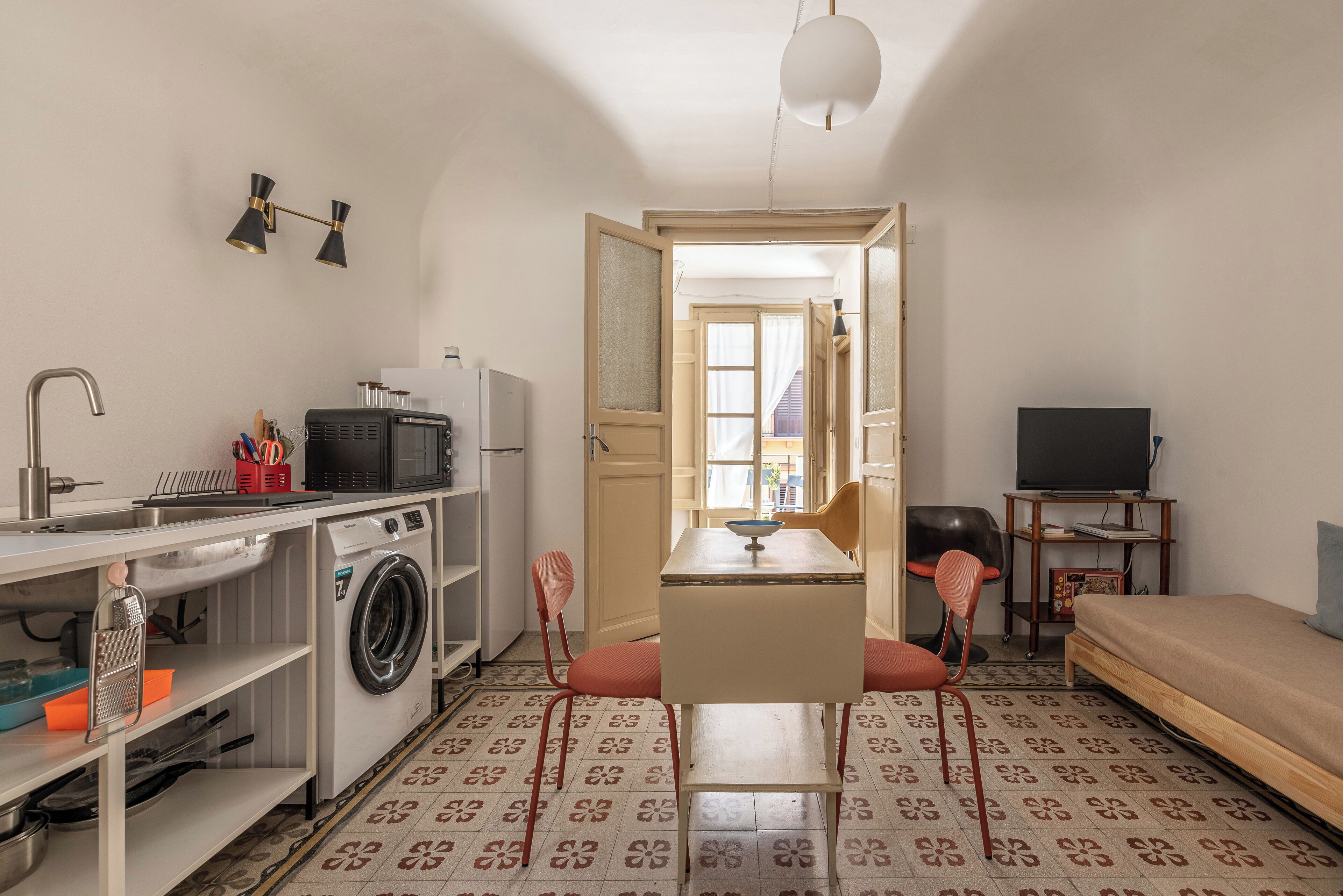 Een kleine kamer met een wasmachine, een wastafel, een klein tafeltje met stoelen, een bed en een televisie.