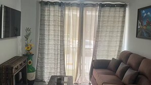 A living room with a brown sofa, a wooden coffee table, a TV mounted on the wall, and a large window with striped curtains.