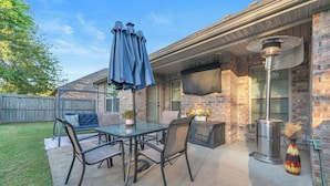 Un patio exterior con una mesa de vidrio, sillas, un paraguas azul y un jarrón decorativo.