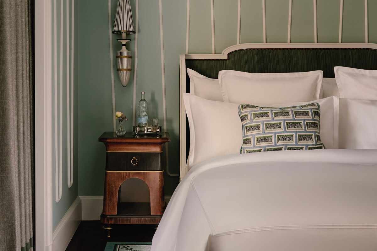 A neatly made bed with white linens and a patterned pillow, a bedside table with a lamp, and a decorative wall panel.