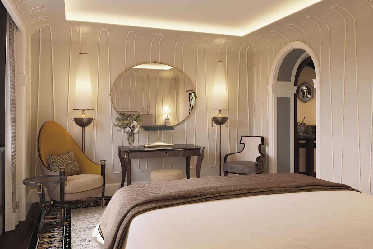 A hotel room with a large bed, a desk, two chairs, a mirror, and a decorative vase.