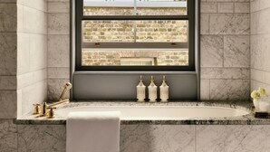 A bathroom with a marble sink, gold fixtures, and a view of a brick wall through a window. - Cambridge House, Auberge Collection (London)