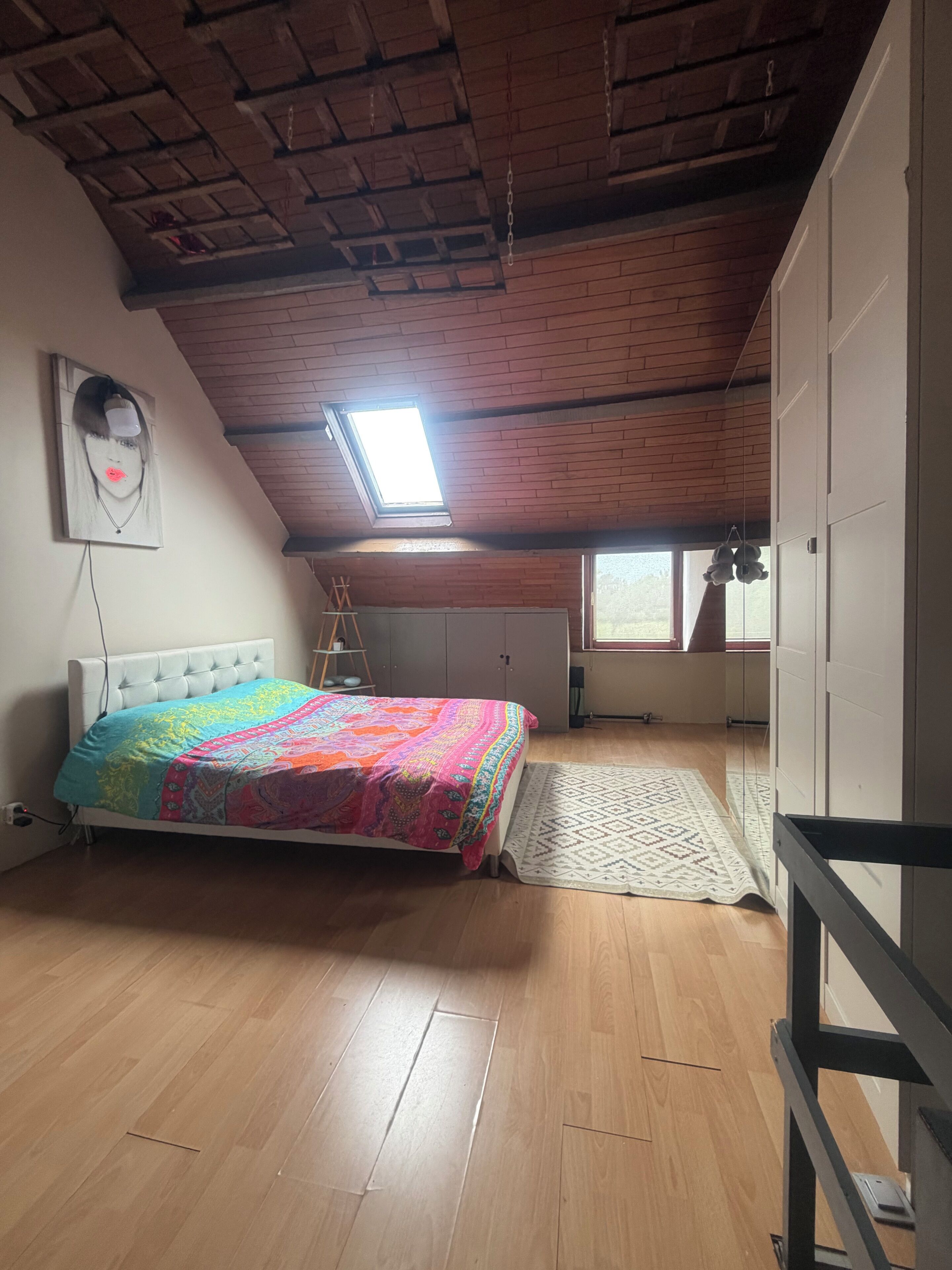 A bedroom with a bed, a skylight, a window, and a wooden ceiling.