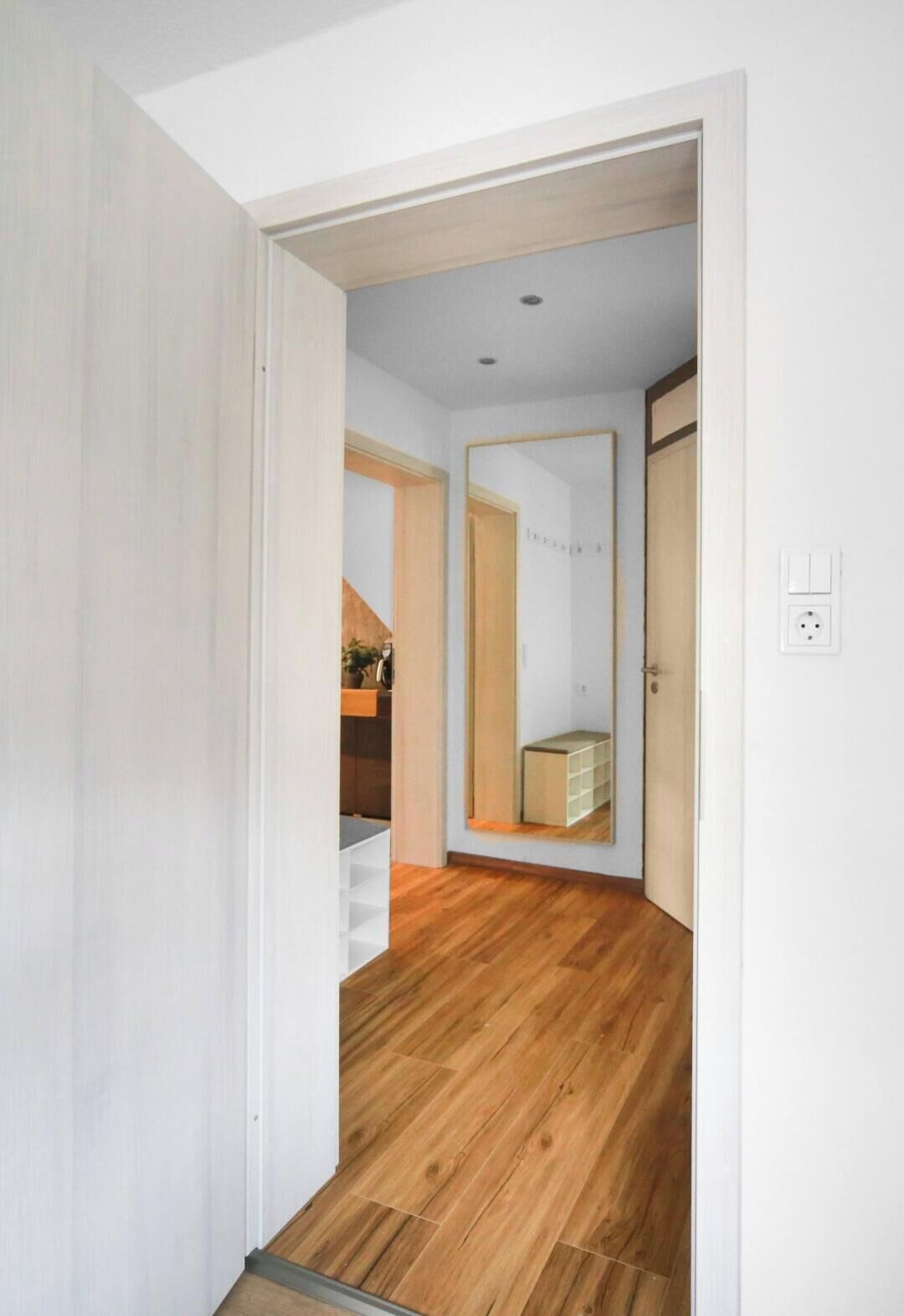Un couloir doté d’un plancher en bois, d’une porte blanche et d’un miroir qui reflète une pièce contenant un lit et une commode.