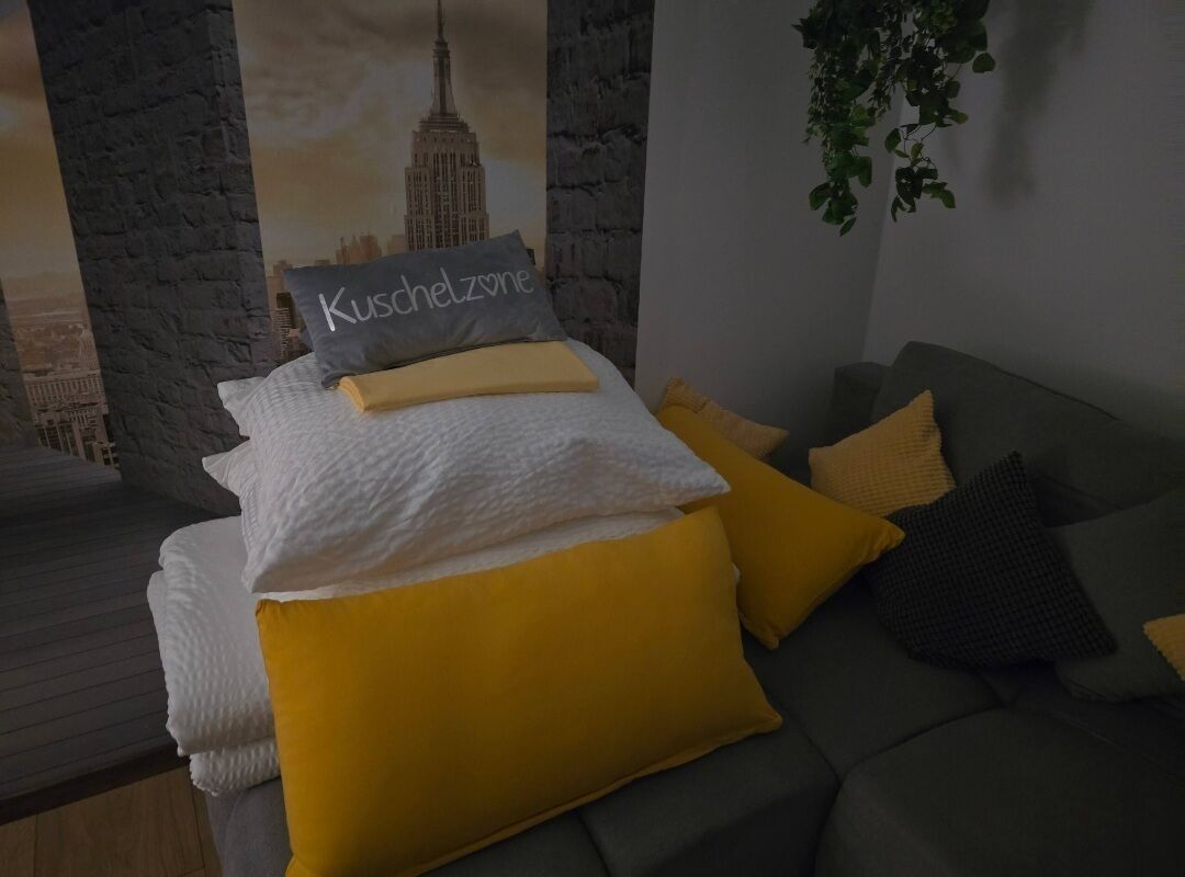 A bed with white and yellow bedding, a pillow with "Kuscheltzone" text, and a grey couch with yellow cushions.