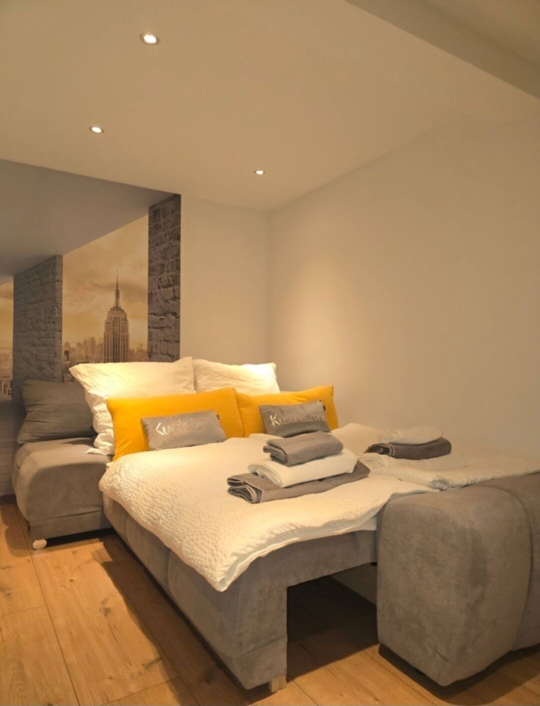 A modern bedroom with a large bed, a sofa, and a cityscape mural on the wall.