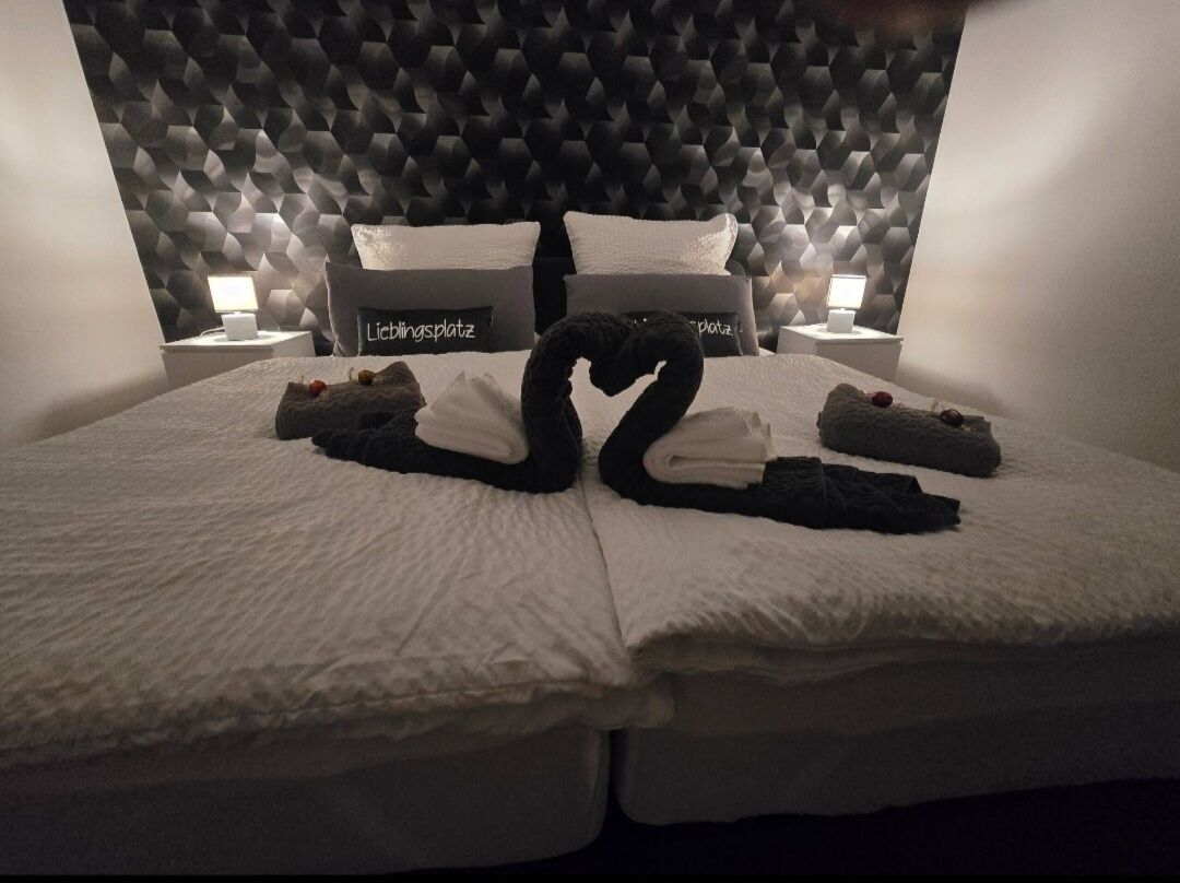 A hotel room with a unique geometric wall design, a neatly made bed with towels arranged in a heart shape, and two pillows with text on them.