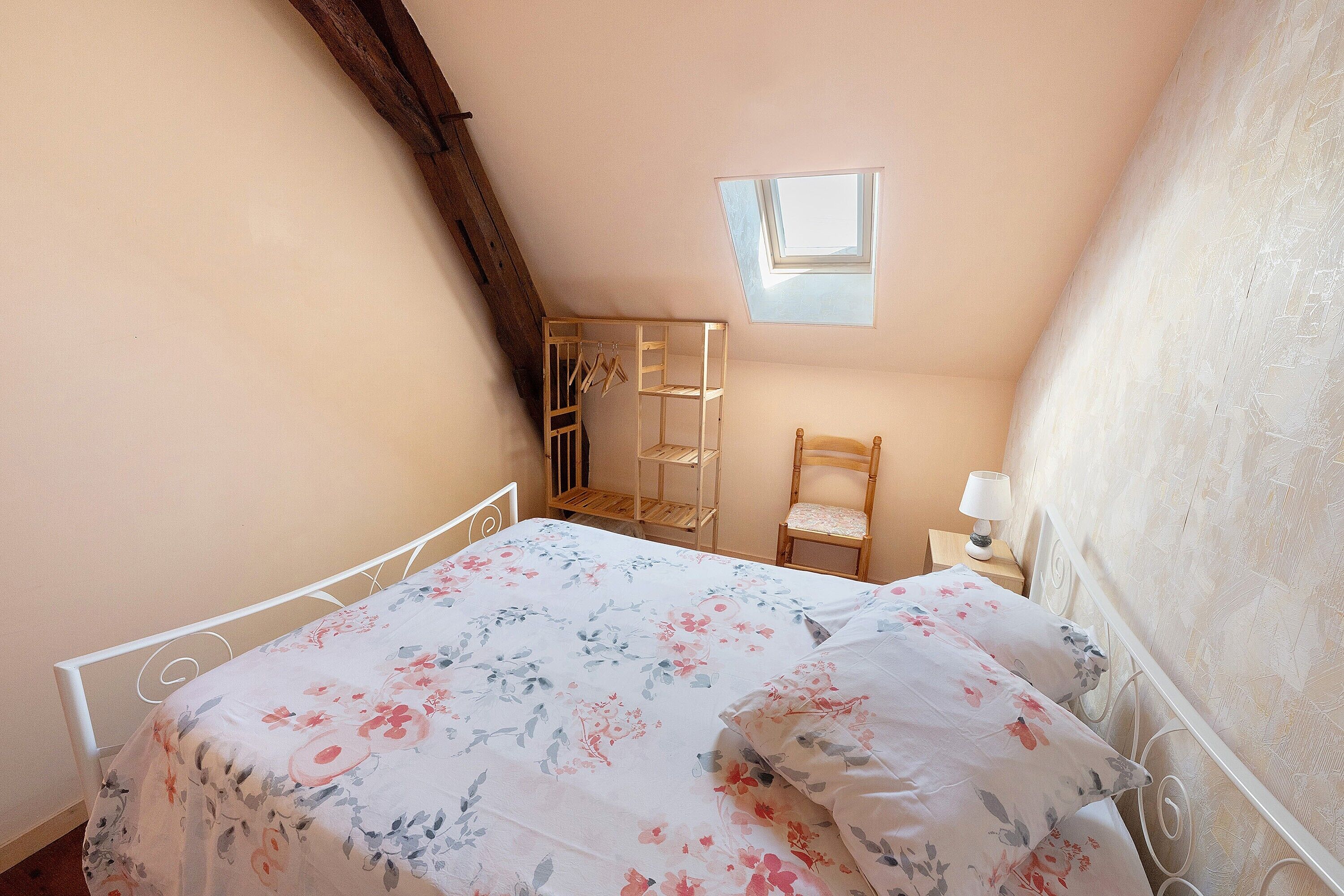 Une chambre avec un plafond en pente, un lit recouvert d’une literie à motifs floraux, une échelle en bois, une chaise et une petite table avec une lampe.
