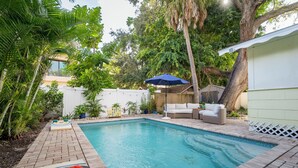 Un patio trasero con piscina, un área de descanso con sofá y mesa, y un espacio de estar techado.