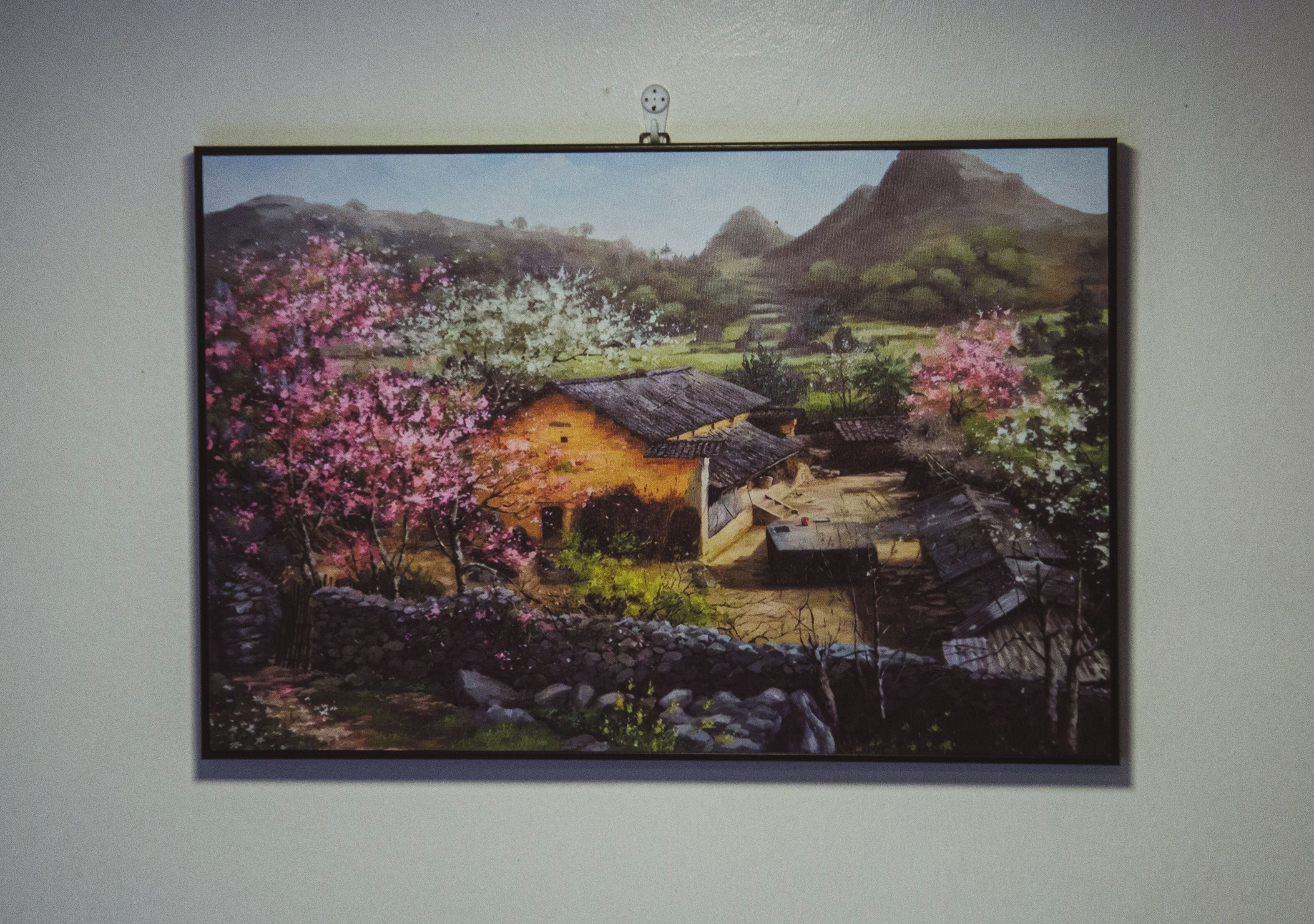 A framed landscape painting of a rural scene with a house, blooming trees, and a mountain in the background.