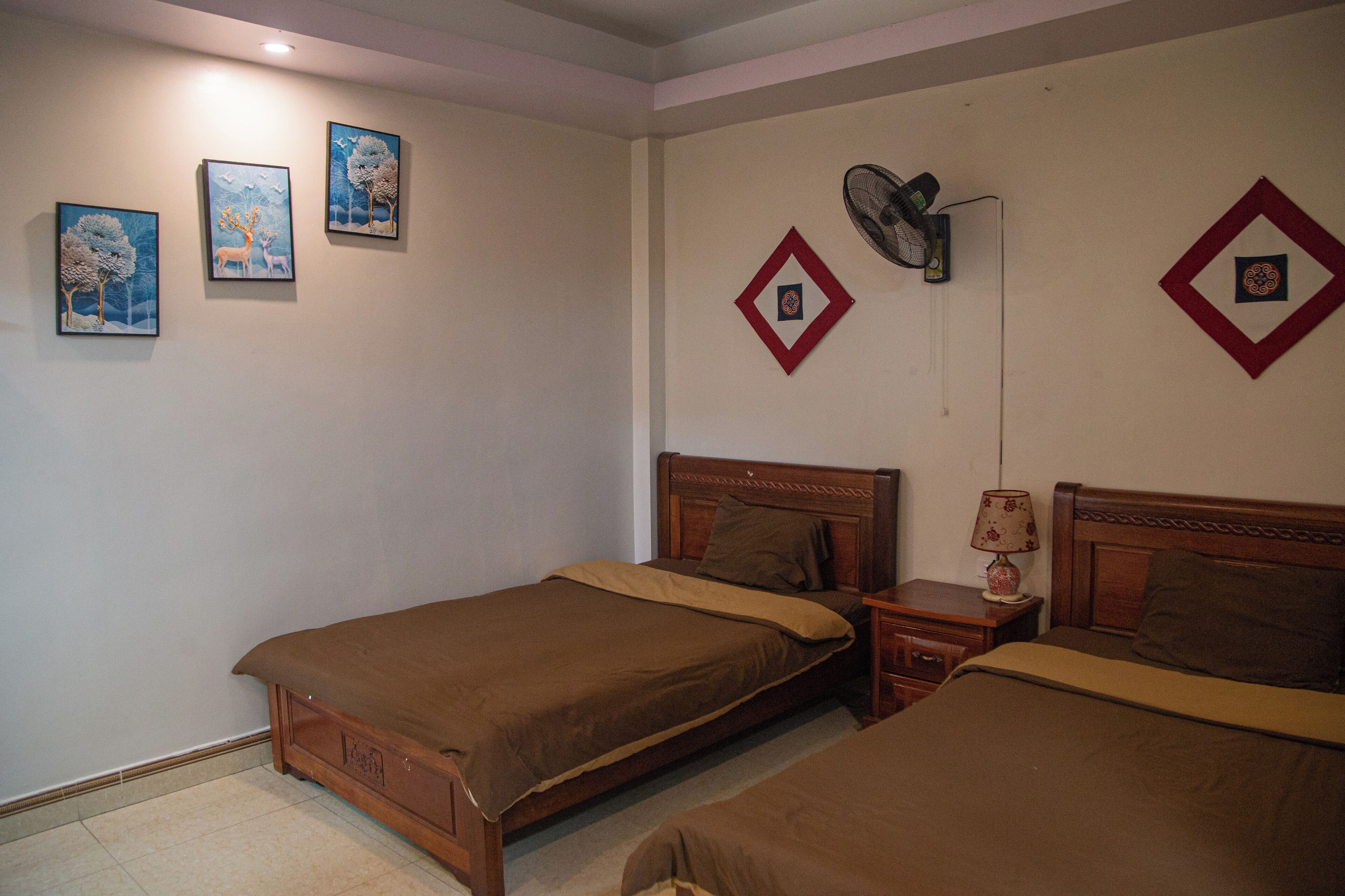 Une chambre avec deux lits simples, une table de chevet avec une lampe, un ventilateur fixé au mur et trois tableaux encadrés accrochés au mur.