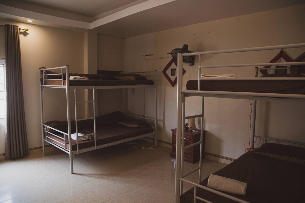 A dormitory room with bunk beds, a small cabinet, and a window with curtains.