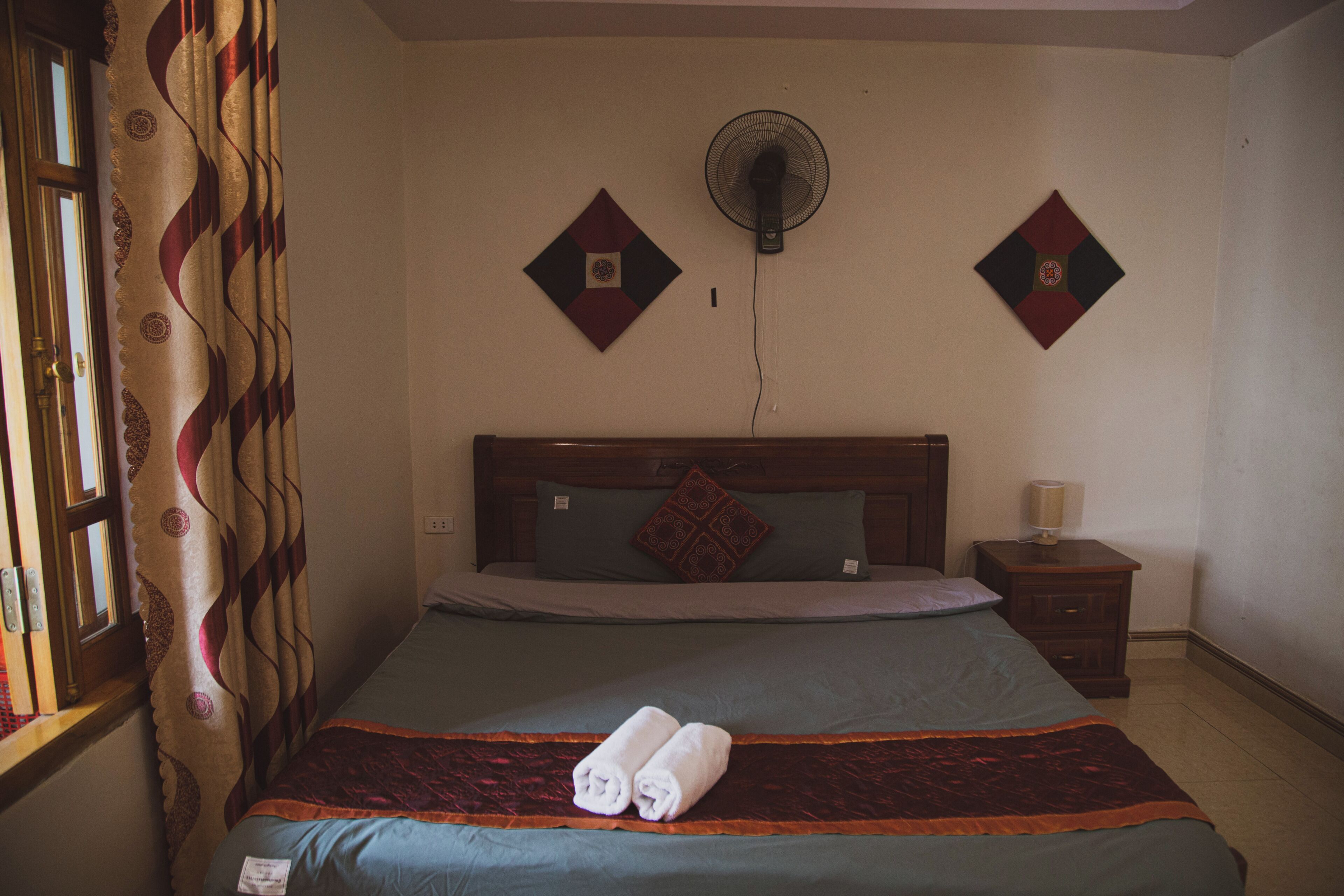 Un dormitorio con cama, mesita de noche, ventilador de pared y ventana con cortinas.