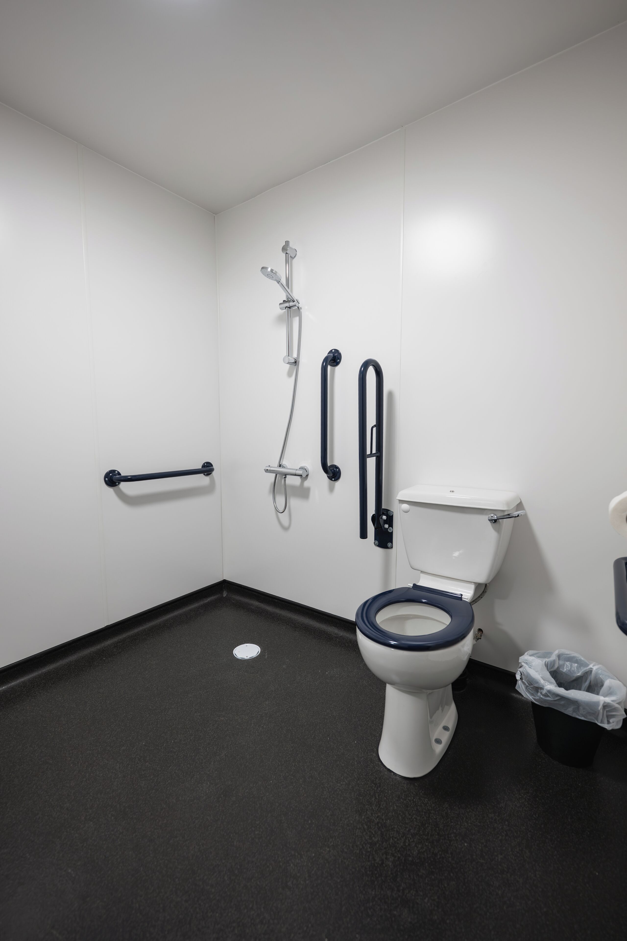 A bathroom with a white toilet, black floor, and white walls. There are grab bars on the wall and a trash can.