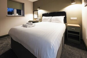 A neatly made bed with white linens, a bedside table with a lamp, and a window with a view of trees.