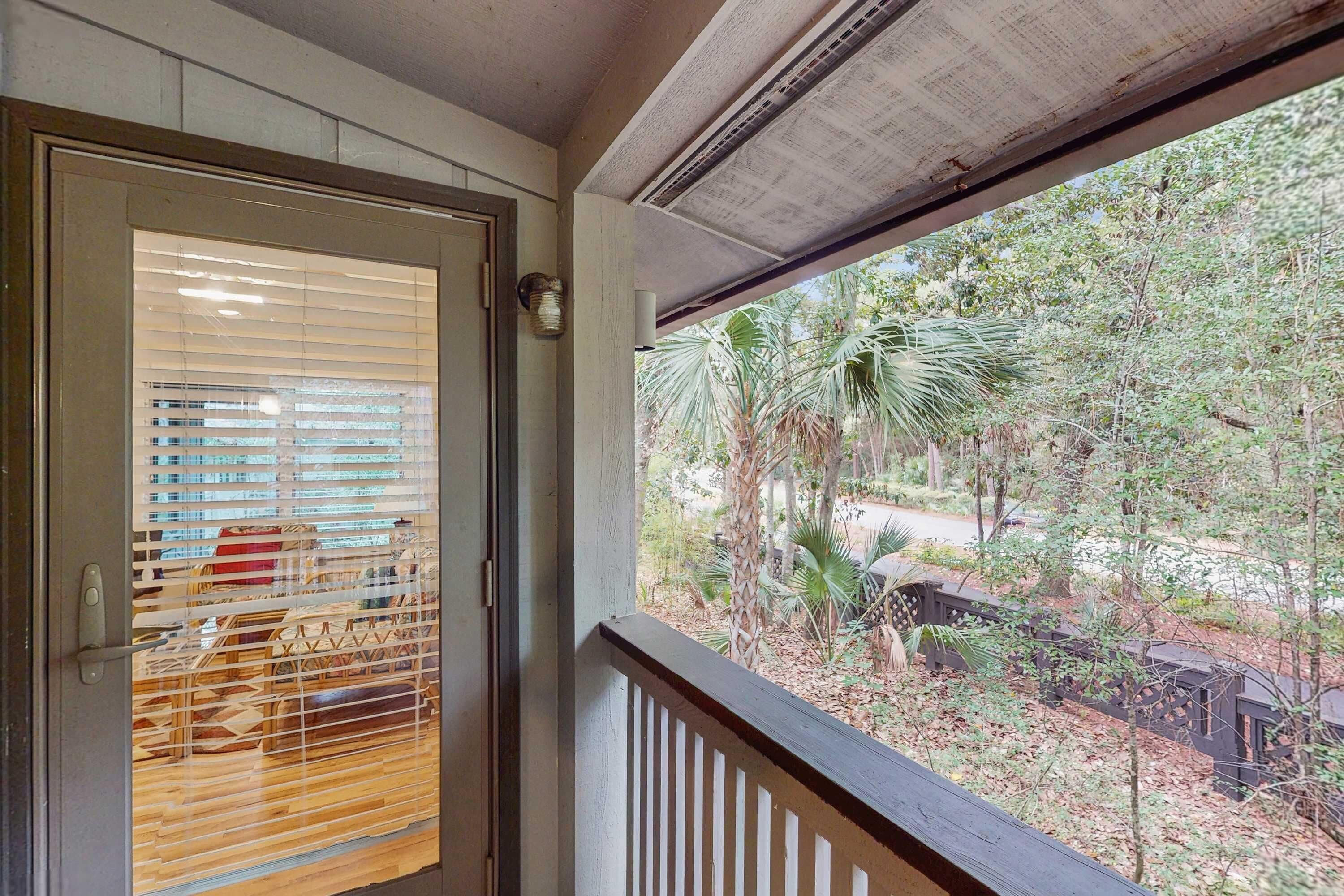 Quarto com porta de madeira, janela com persianas e vista para um jardim com árvores e um caminho.