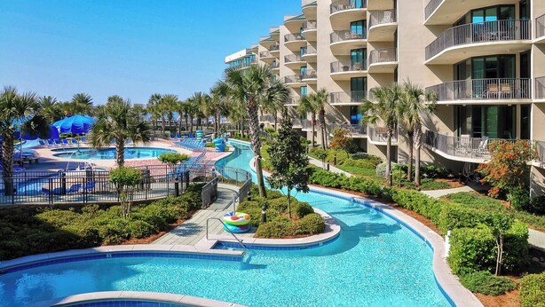 A resort with a large swimming pool, palm trees, and a multi-story building with balconies.