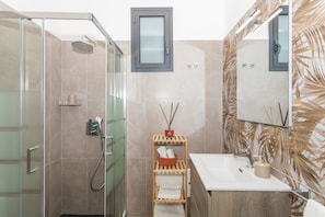 A modern bathroom with a glass-enclosed shower, a wooden shelf with toiletries, a sink with a mirror, and a decorative wall with leaf patterns.