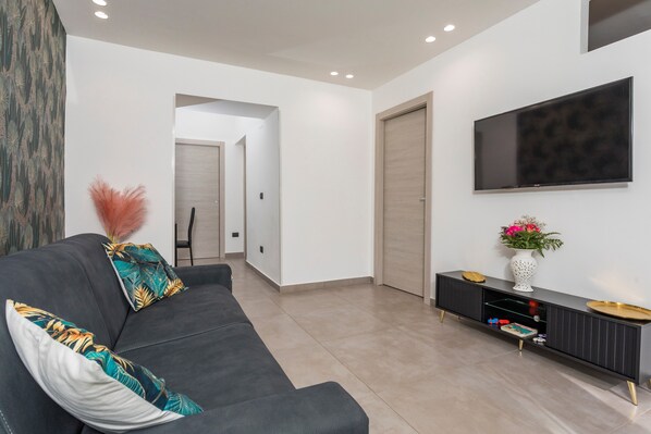 A modern living room with a grey sofa, a TV mounted on the wall, a wooden door, and a small side table with a vase.