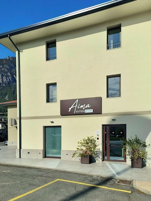 Um edifício com uma placa indicando 'Aima Boutique Rooms' e uma porta com painel de vidro. Há duas plantas em vasos na calçada.