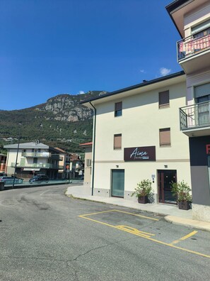 A building with a sign for 'Aina Boutique Rooms' on a street with a mountain in the background. - Aima Boutique Rooms (Costa Volpino)