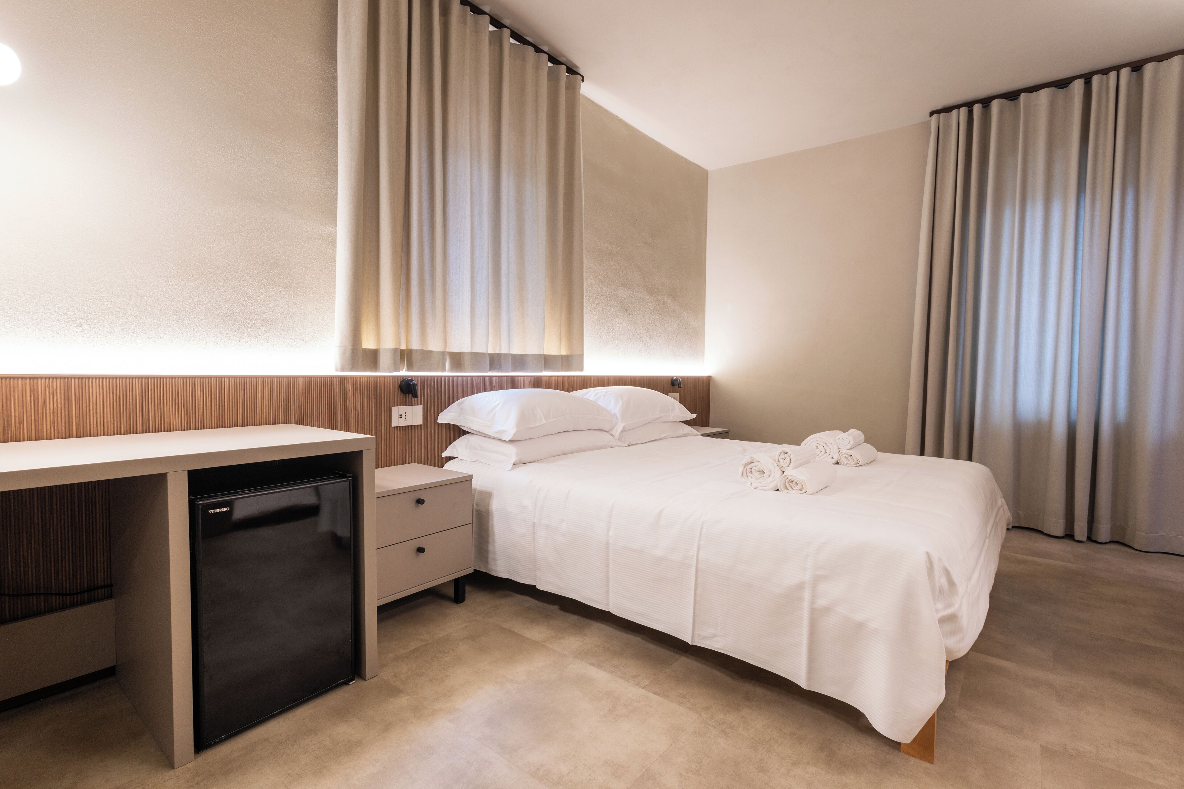 Habitación de hotel con cama, mesitas de noche, escritorio con refrigerador y cortinas en las ventanas.