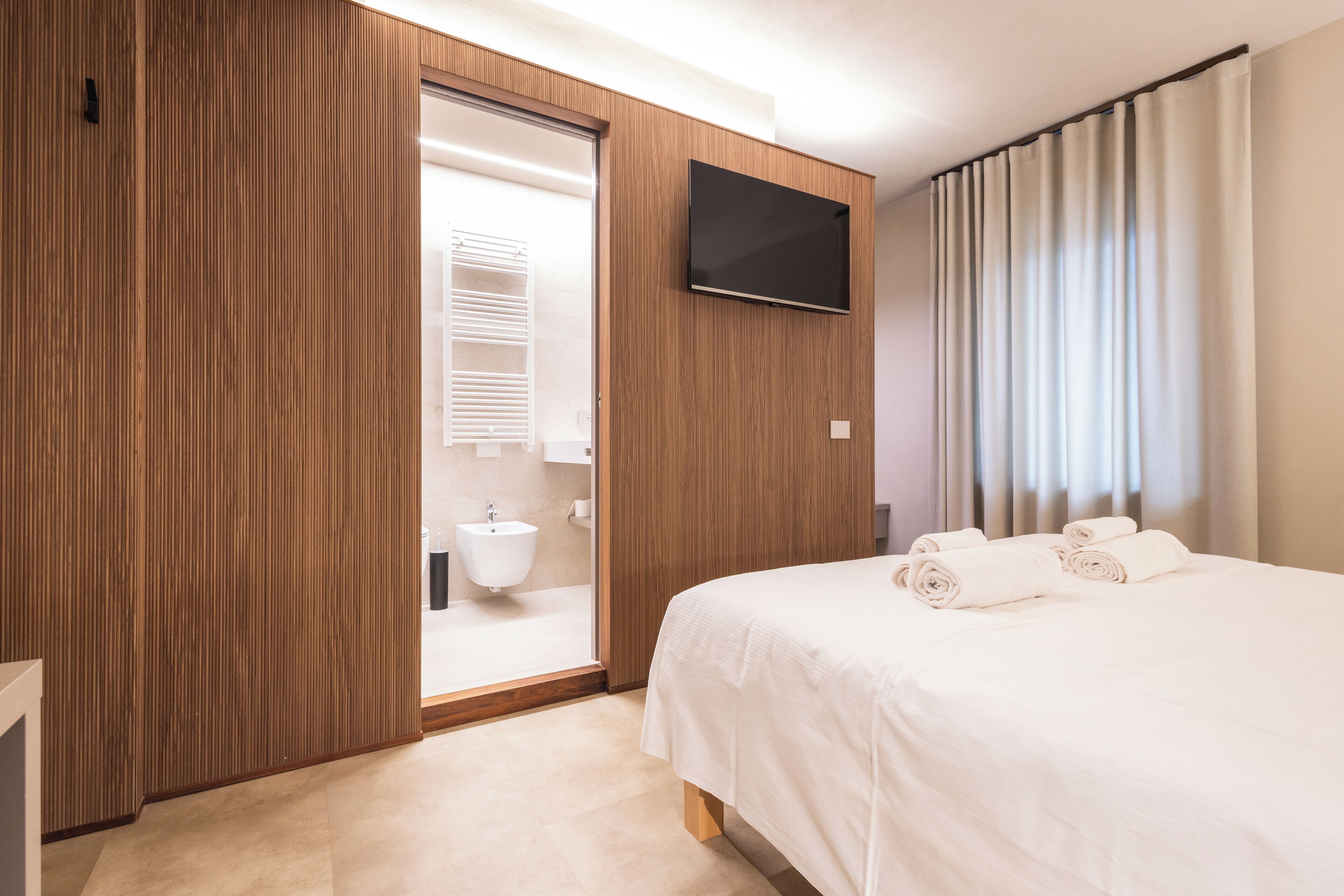 Una habitación de hotel con un armario de madera, un televisor de pantalla plana, una cama con ropa blanca y un baño con lavabo e inodoro.