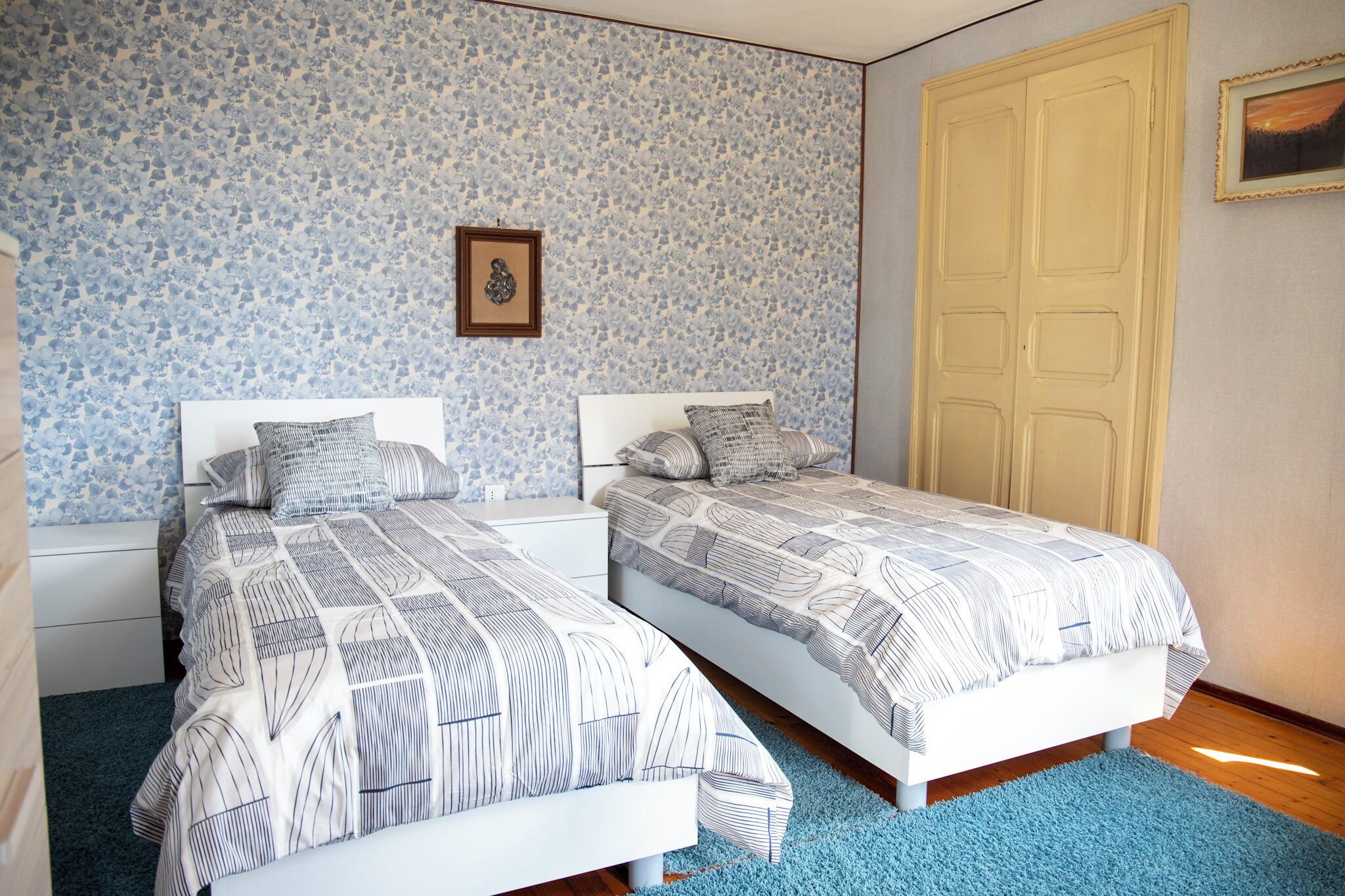 A bedroom with two beds, a blue floral wallpaper, a framed picture on the wall, and a wooden door.