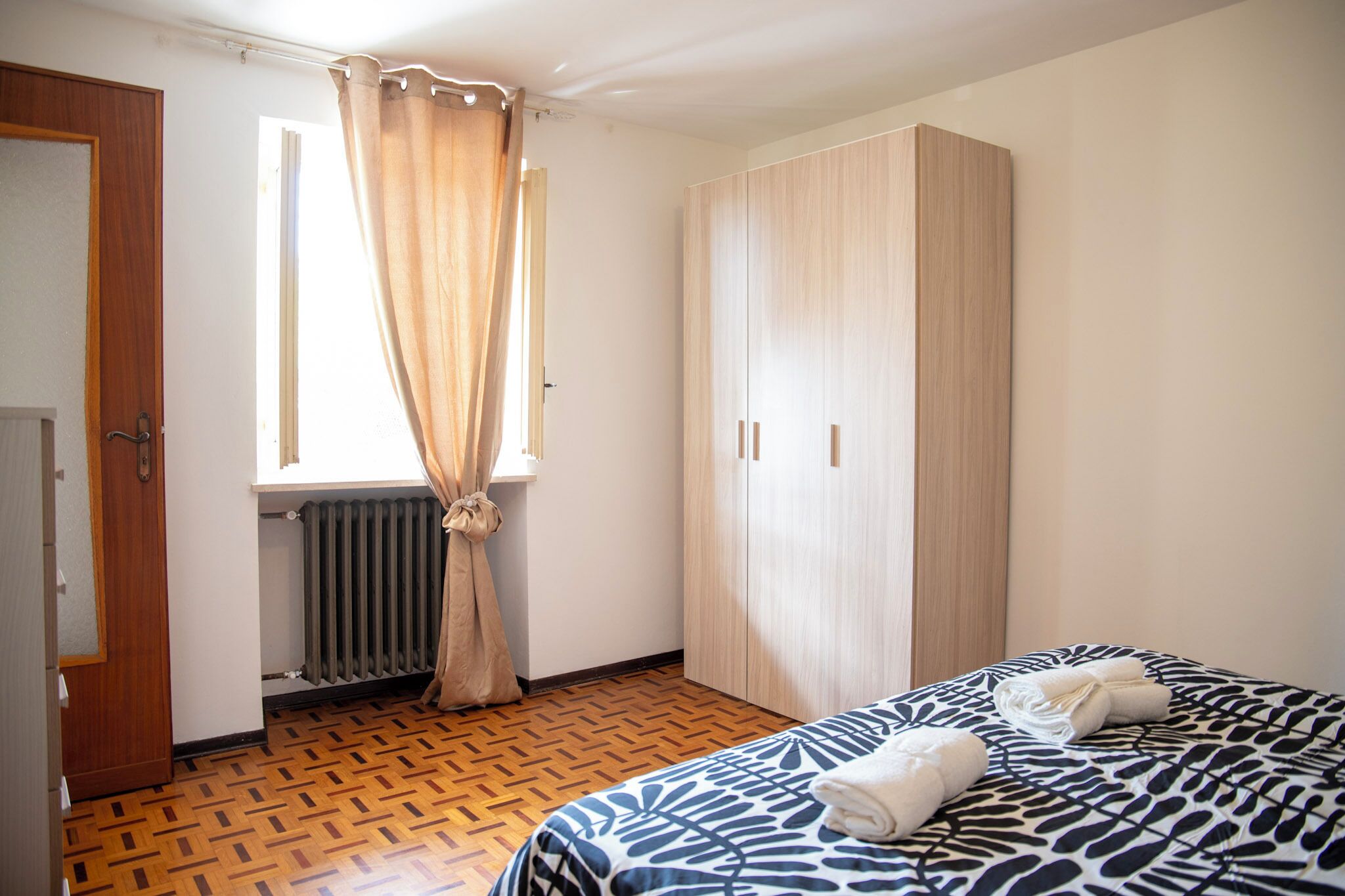 Ein Schlafzimmer mit Bett, Kleiderschrank, Heizkörper und einem Fenster mit Vorhängen.