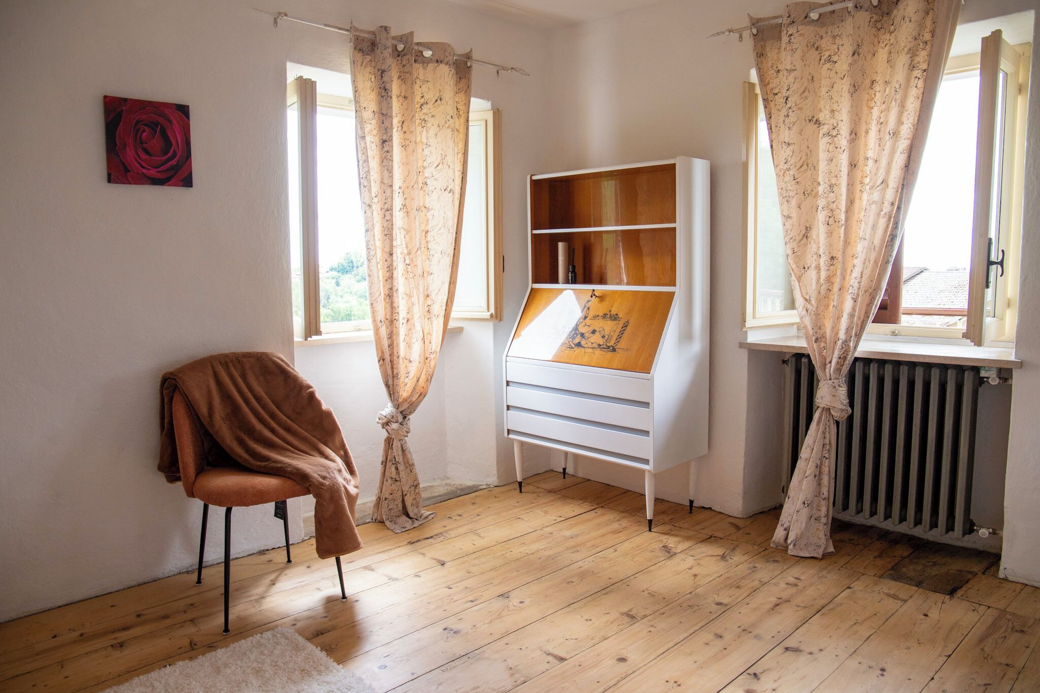A room with wooden flooring, a white cabinet, a brown chair, and two windows with floral curtains.