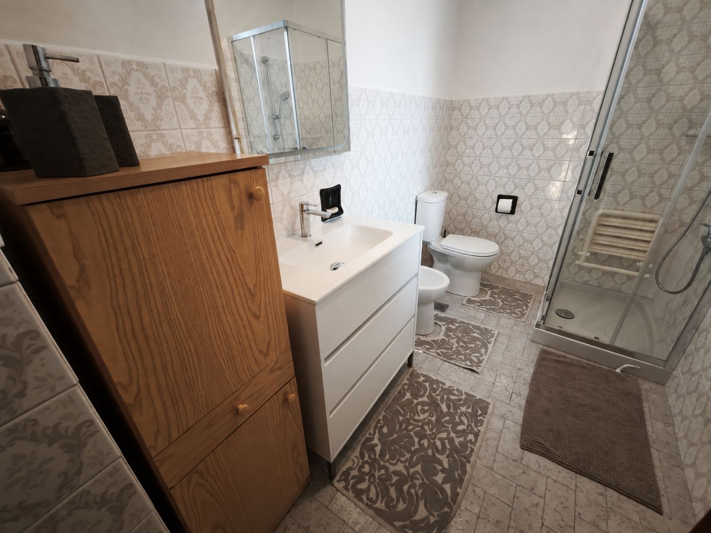 Un baño con tocador de madera, un lavamanos blanco, un inodoro y una zona de ducha con mampara de vidrio.